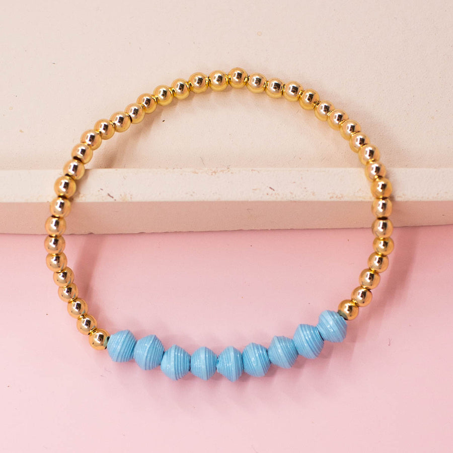 stackable bracelet made of 14k gold plated brass beads and handmade paper beads. The brass beads are round and shiny, while the paper beads are colorful and textured, creating a striking contrast. The bracelet is designed to be worn with other bracelets and features an elastic band that fits most wrist sizes. It is displayed on a neutral background with good lighting that highlights the details and craftsmanship of the bracelet.
