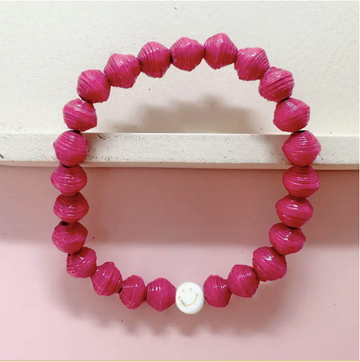 A close-up photo of an ethically made bracelet made of bright pink, handmade paper beads and a smiley face charm