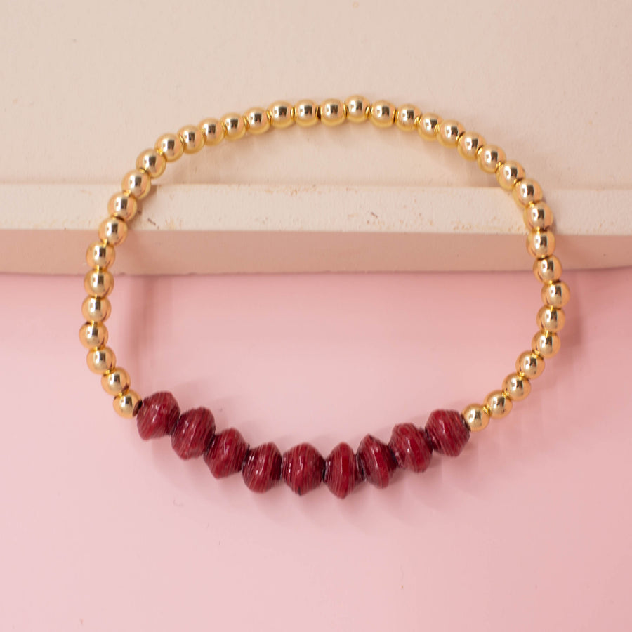 stackable bracelet made of 14k gold plated brass beads and handmade paper beads. The brass beads are round and shiny, while the paper beads are colorful and textured, creating a striking contrast. The bracelet is designed to be worn with other bracelets and features an elastic band that fits most wrist sizes. It is displayed on a neutral background with good lighting that highlights the details and craftsmanship of the bracelet.