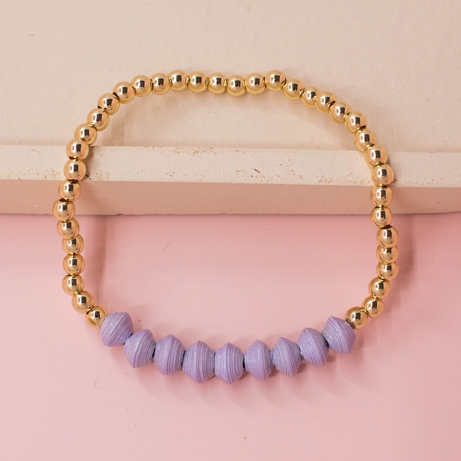 stackable bracelet made of 14k gold plated brass beads and handmade paper beads. The brass beads are round and shiny, while the paper beads are colorful and textured, creating a striking contrast. The bracelet is designed to be worn with other bracelets and features an elastic band that fits most wrist sizes. It is displayed on a neutral background with good lighting that highlights the details and craftsmanship of the bracelet.