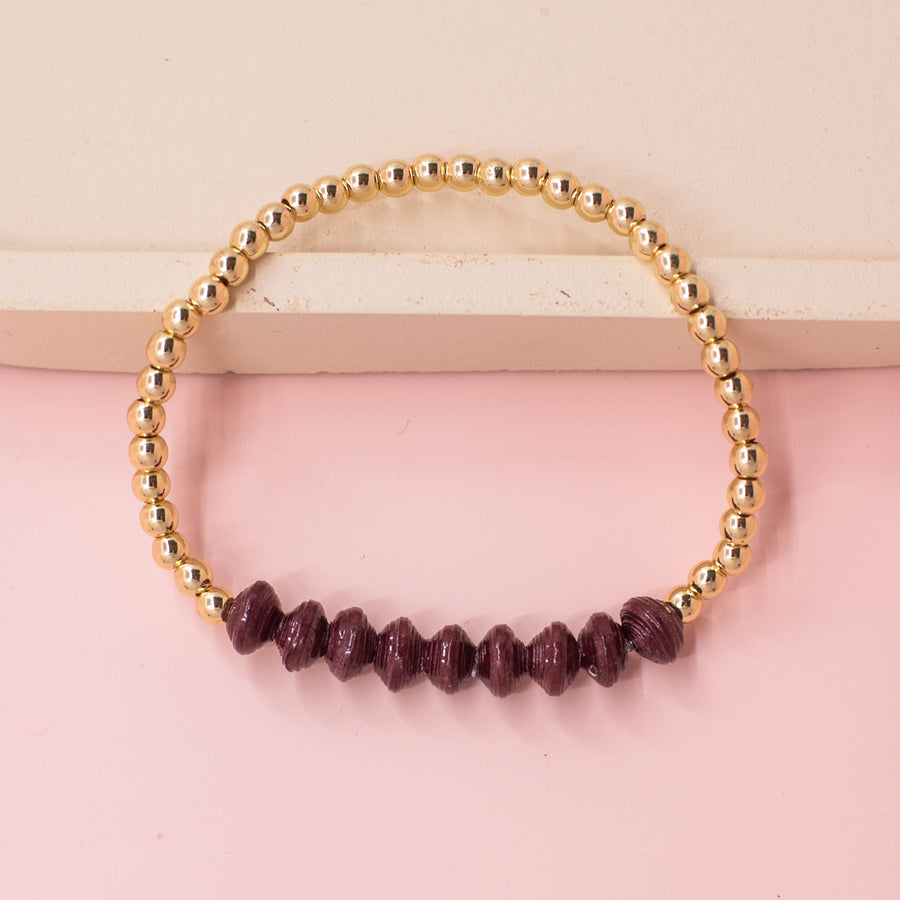 stackable bracelet made of 14k gold plated brass beads and handmade paper beads. The brass beads are round and shiny, while the paper beads are colorful and textured, creating a striking contrast. The bracelet is designed to be worn with other bracelets and features an elastic band that fits most wrist sizes. It is displayed on a neutral background with good lighting that highlights the details and craftsmanship of the bracelet.