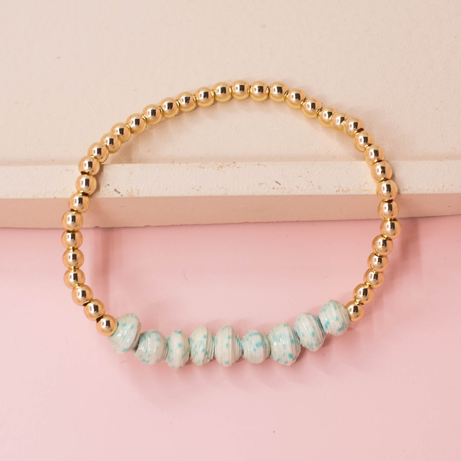 stackable bracelet made of 14k gold plated brass beads and handmade paper beads. The brass beads are round and shiny, while the paper beads are colorful and textured, creating a striking contrast. The bracelet is designed to be worn with other bracelets and features an elastic band that fits most wrist sizes. It is displayed on a neutral background with good lighting that highlights the details and craftsmanship of the bracelet.