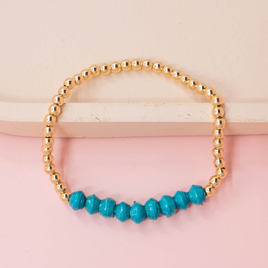 stackable bracelet made of 14k gold plated brass beads and handmade paper beads. The brass beads are round and shiny, while the paper beads are colorful and textured, creating a striking contrast. The bracelet is designed to be worn with other bracelets and features an elastic band that fits most wrist sizes. It is displayed on a neutral background with good lighting that highlights the details and craftsmanship of the bracelet.