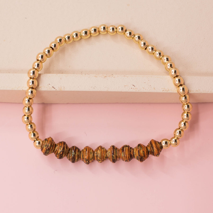 stackable bracelet made of 14k gold plated brass beads and handmade paper beads. The brass beads are round and shiny, while the paper beads are colorful and textured, creating a striking contrast. The bracelet is designed to be worn with other bracelets and features an elastic band that fits most wrist sizes. It is displayed on a neutral background with good lighting that highlights the details and craftsmanship of the bracelet.