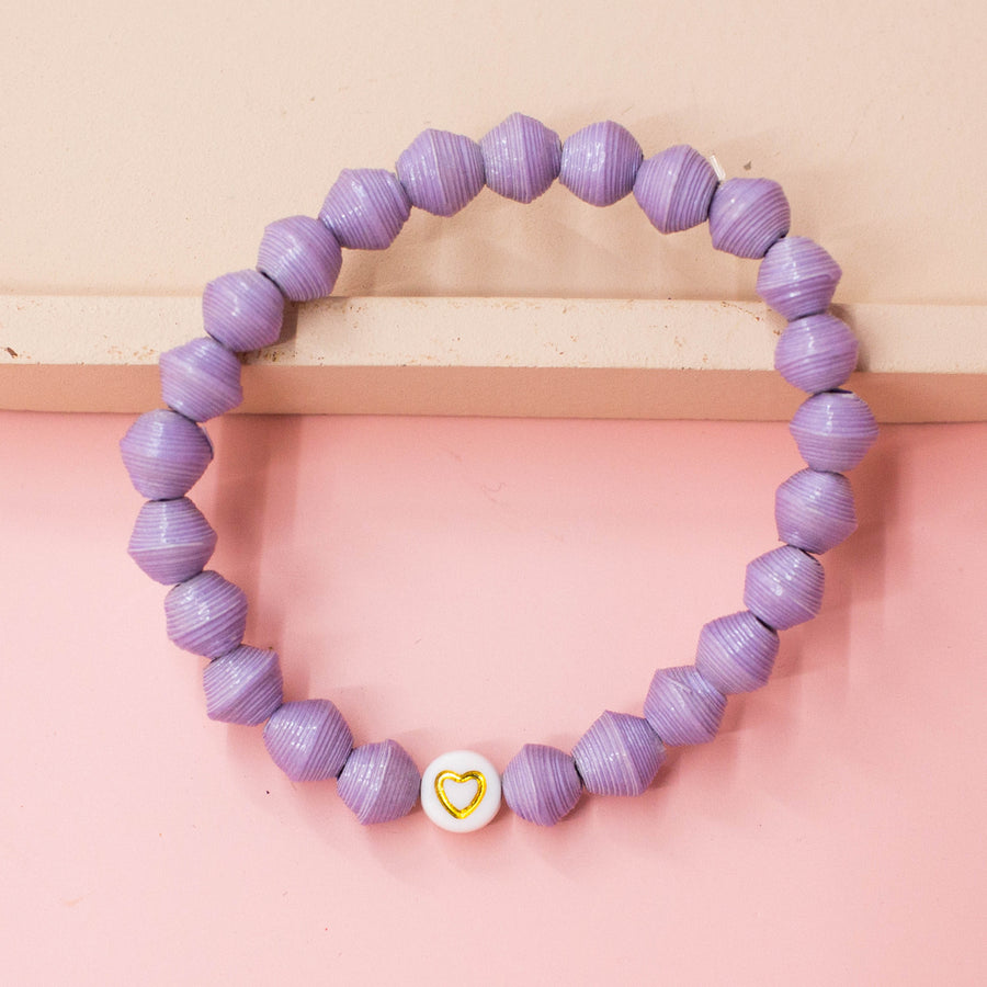 A close-up photo of an ethically made bracelet made of light purple, handmade paper beads and a heart symbol bead