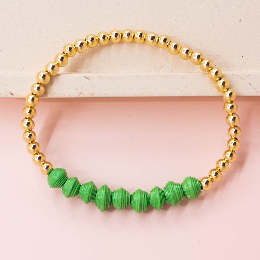 stackable bracelet made of 14k gold plated brass beads and handmade paper beads. The brass beads are round and shiny, while the paper beads are colorful and textured, creating a striking contrast. The bracelet is designed to be worn with other bracelets and features an elastic band that fits most wrist sizes. It is displayed on a neutral background with good lighting that highlights the details and craftsmanship of the bracelet.