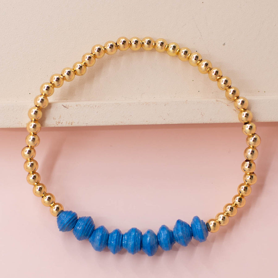 stackable bracelet made of 14k gold plated brass beads and handmade paper beads. The brass beads are round and shiny, while the paper beads are colorful and textured, creating a striking contrast. The bracelet is designed to be worn with other bracelets and features an elastic band that fits most wrist sizes. It is displayed on a neutral background with good lighting that highlights the details and craftsmanship of the bracelet.